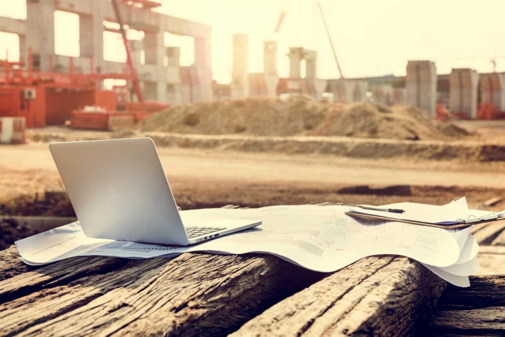 Open silver laptop and white blueprint papers on a weathered wooden surface at a sunny construction site with cranes, containers, and piles of dirt in the background.