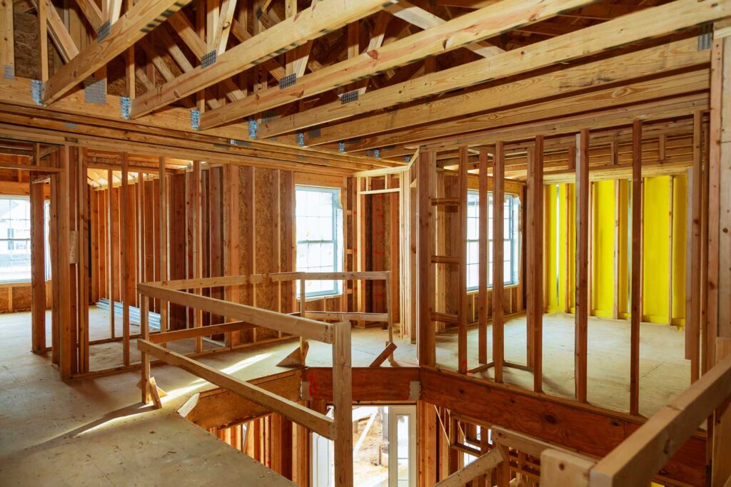 Interior view of a house under construction showcasing wooden stud framing, plywood flooring, and yellow insulation panels on the wall. A wooden railing is visible in the foreground with a window and door opening in the background.