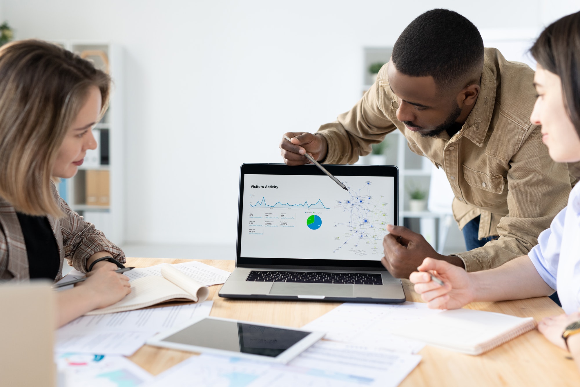 Team of professionals in a meeting, analyzing data on a laptop displaying a graph and pie chart, surrounded by papers and a tablet on a wooden table, with a whiteboard and window in the background.